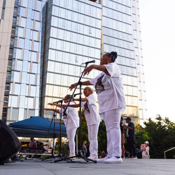 Three musicians in white attire performing outdoors with a microphone and violin, against a backdrop of modern glass buildings bathed in the warm glow of evening light.