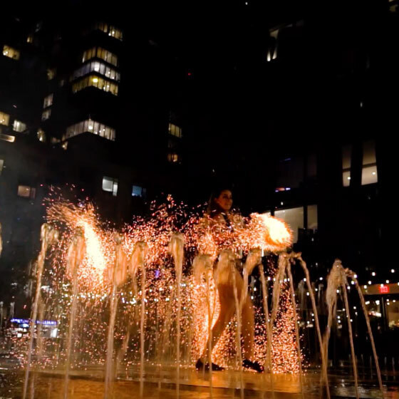 A person standing amidst illuminated water fountains at night, creating a silhouette against a backdrop of city lights.