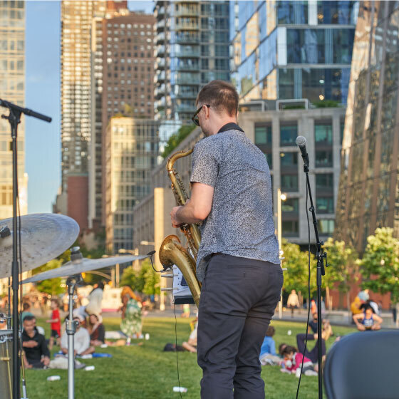 A saxophonist playing at an outdoor event against a backdrop of urban buildings, with an audience relaxed on the grass.