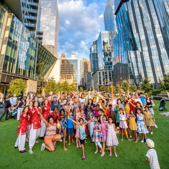 A vibrant gathering of people of different ages and ethnicities celebrating together outdoors in an urban park with skyscrapers in the background.