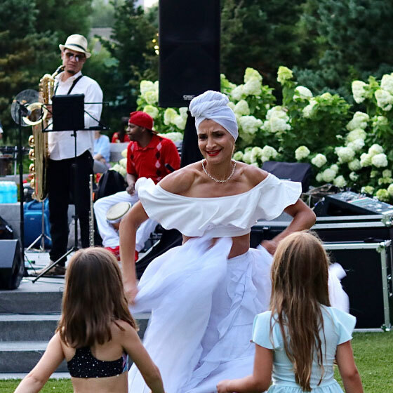 A performer in a white dress and head wrap dances joyfully in the foreground at an outdoor event while musicians play in the background and children watch closely.