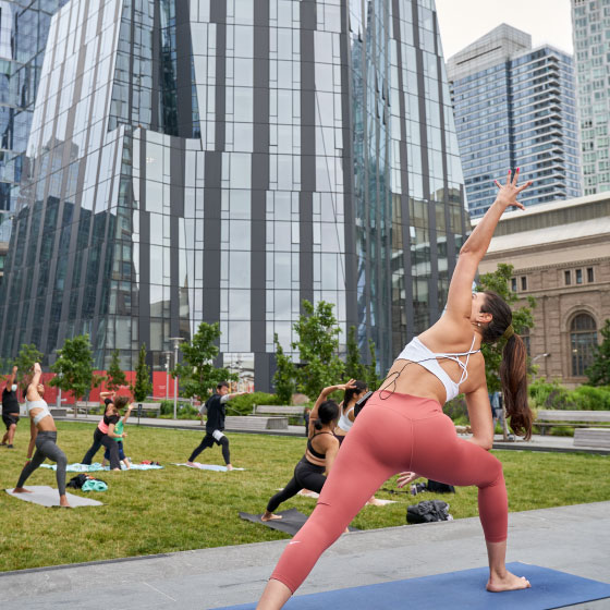 Outdoor yoga class in an urban park with a woman in the foreground performing a yoga pose, surrounded by modern skyscrapers.
