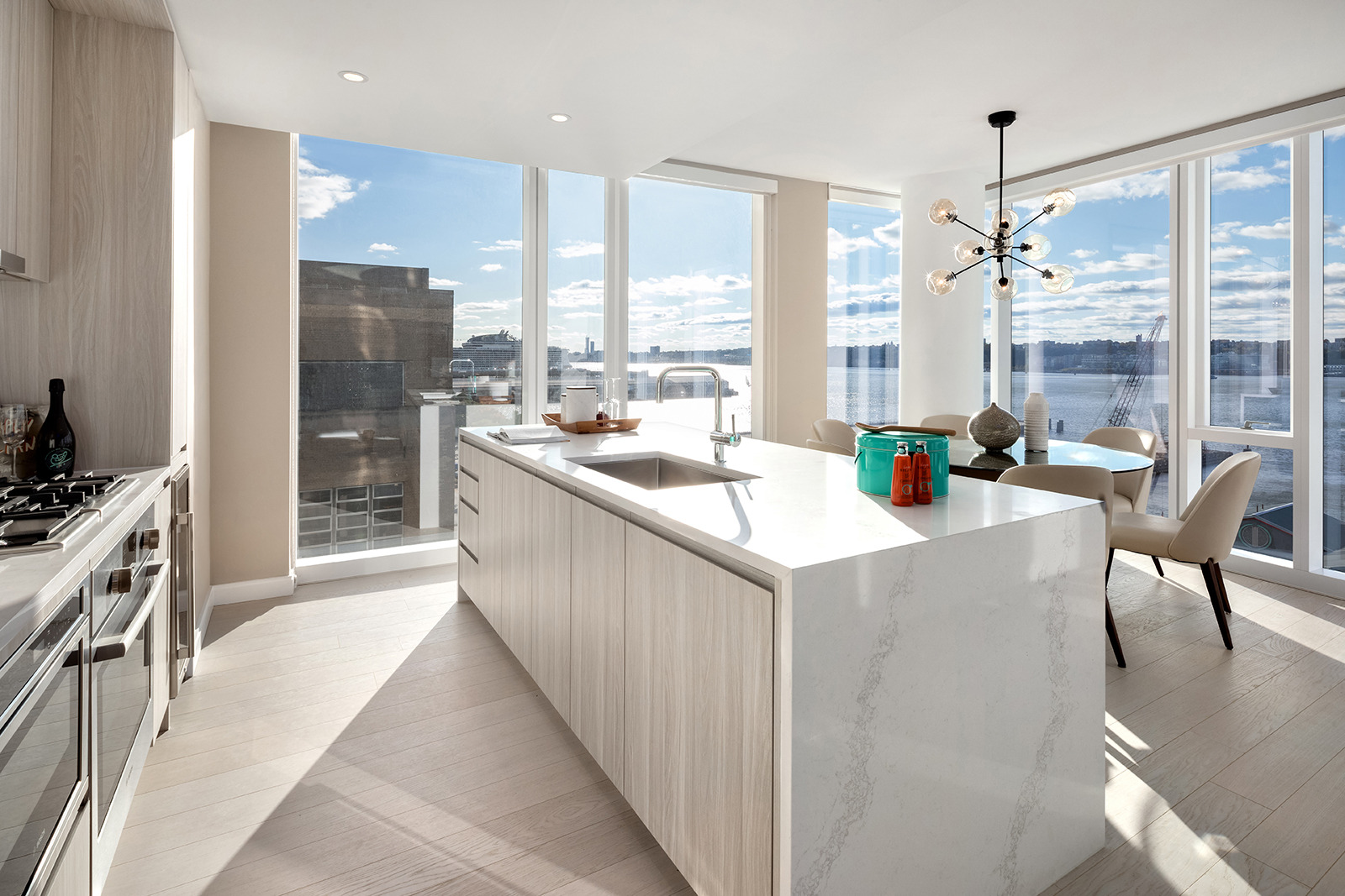 A bright, modern kitchen bathed in natural light with sleek white cabinetry and countertops, featuring a spacious island and an elegant dining area, all overlooking a scenic waterside view.