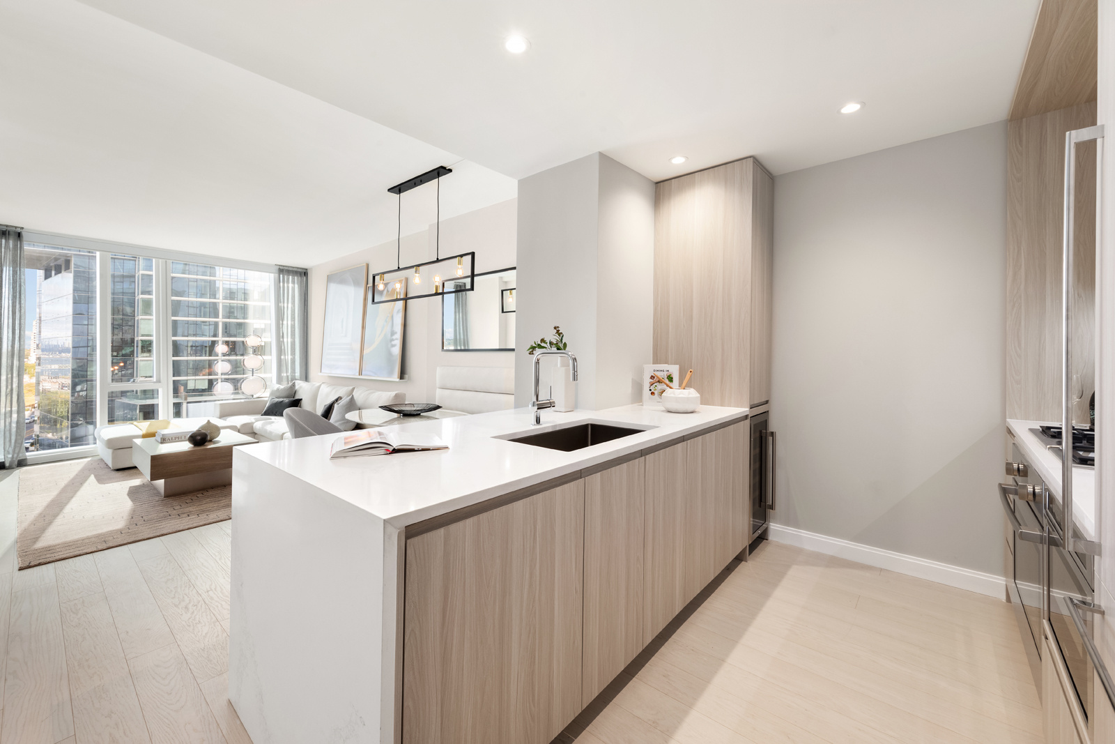 Modern and spacious kitchen with light wood cabinets and countertops, featuring a large island with a sink and a bright living room area in the background, complemented by large windows offering a view of the cityscape.
