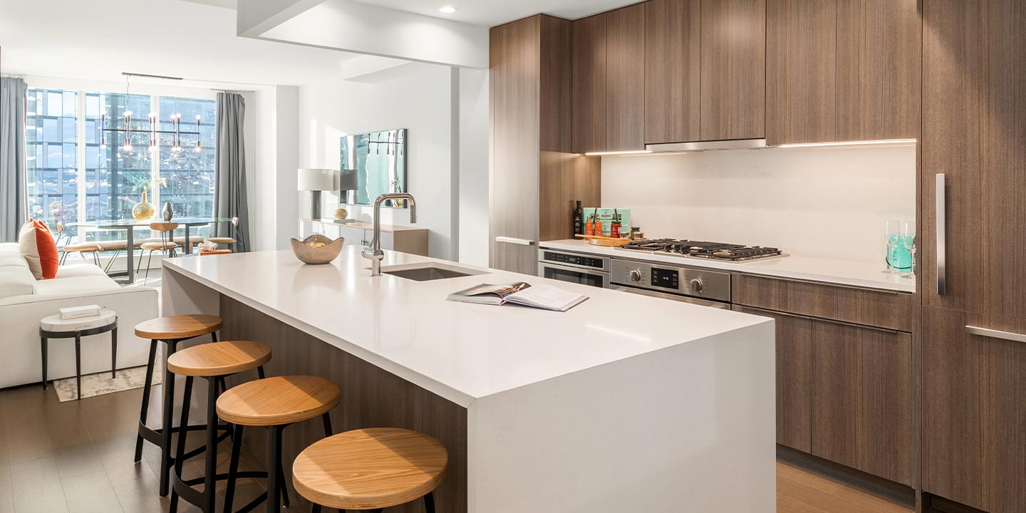 Modern kitchen interior with a sleek white countertop island, wooden cabinetry, and a stylish living area visible in the background, conveying a contemporary urban living space.