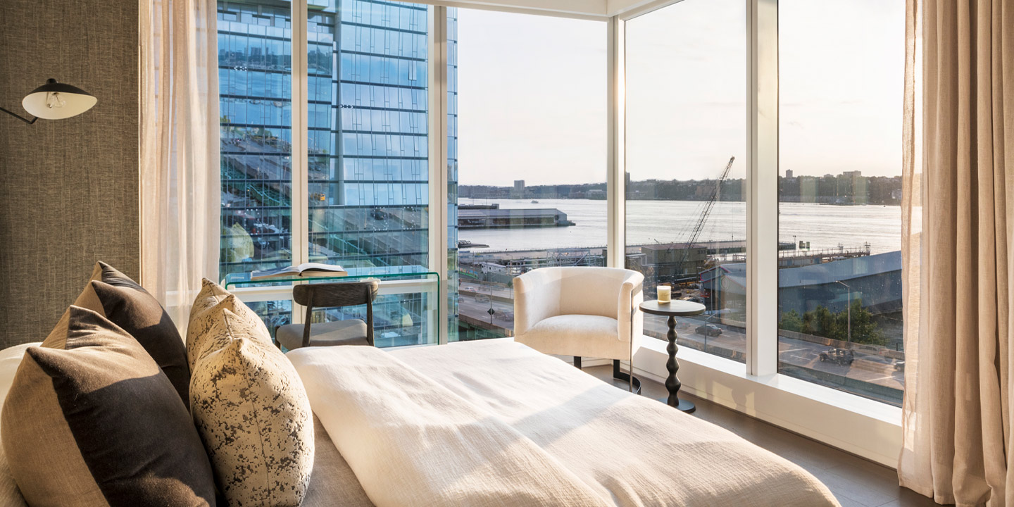 Modern bedroom with a sweeping view of the waterfront, featuring floor-to-ceiling windows that bathe the room in natural light, a cozy bed with decorative pillows, and elegant furnishing that provides a comfortable urban oasis.