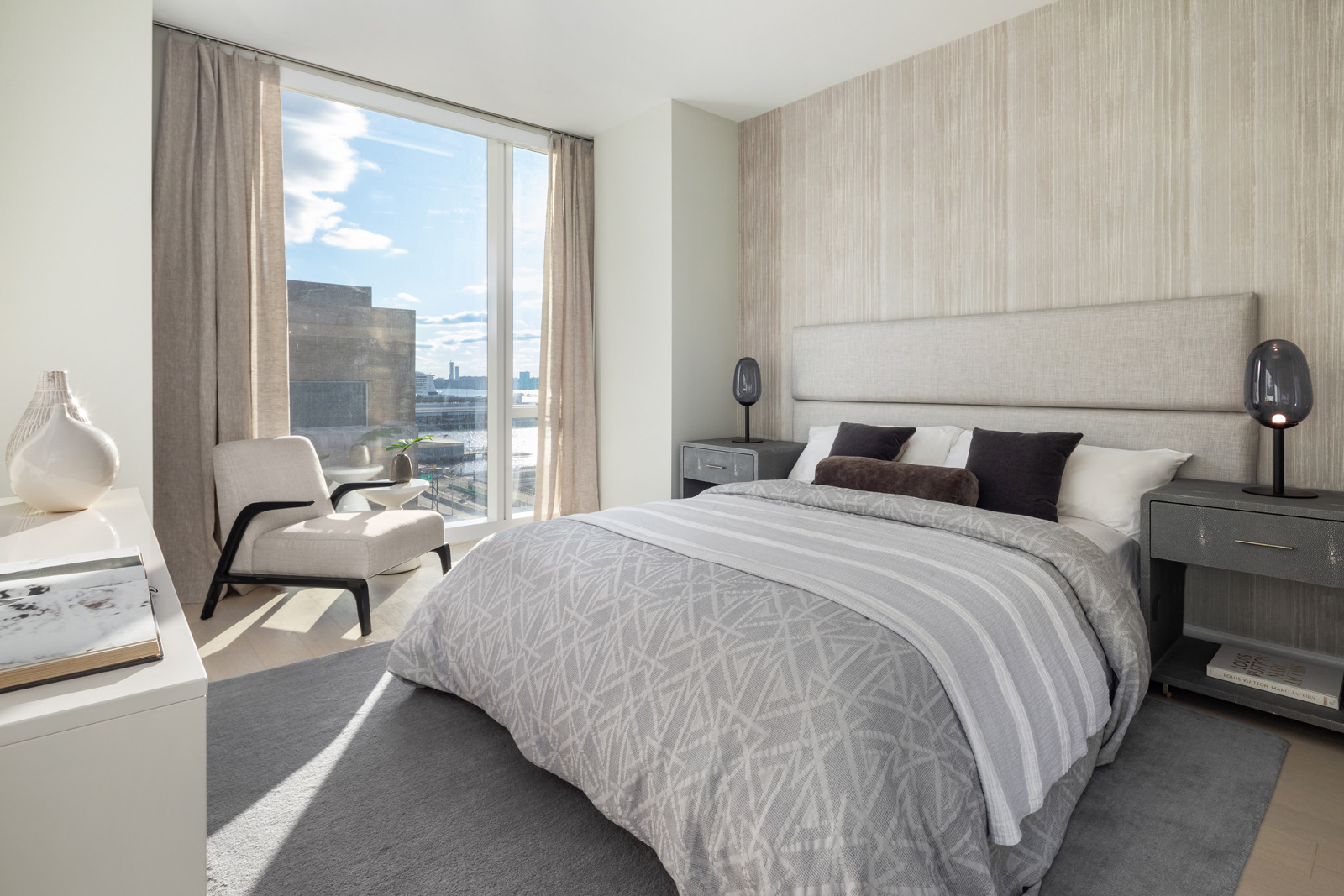 A modern, well-lit bedroom with a large window offering a city view, featuring a neatly made bed with gray and white bedding, flanked by contemporary bedside tables and lamps, with a cozy sitting area in the corner.