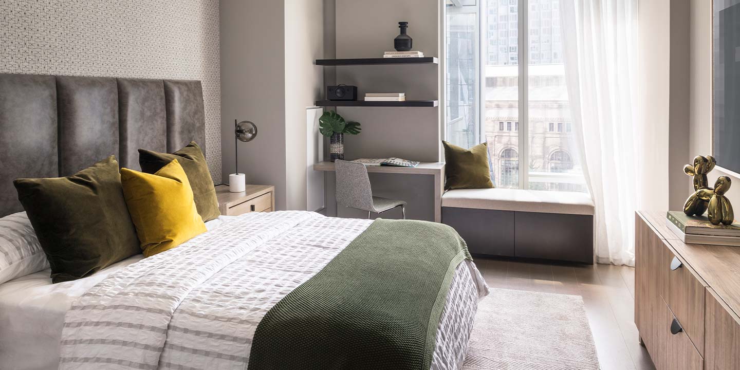 A modern and cozy bedroom with a large window providing ample natural light, featuring a plush gray headboard, green and yellow accent pillows, a muted green throw blanket, and stylish decorative elements, creating an inviting and sophisticated space.
