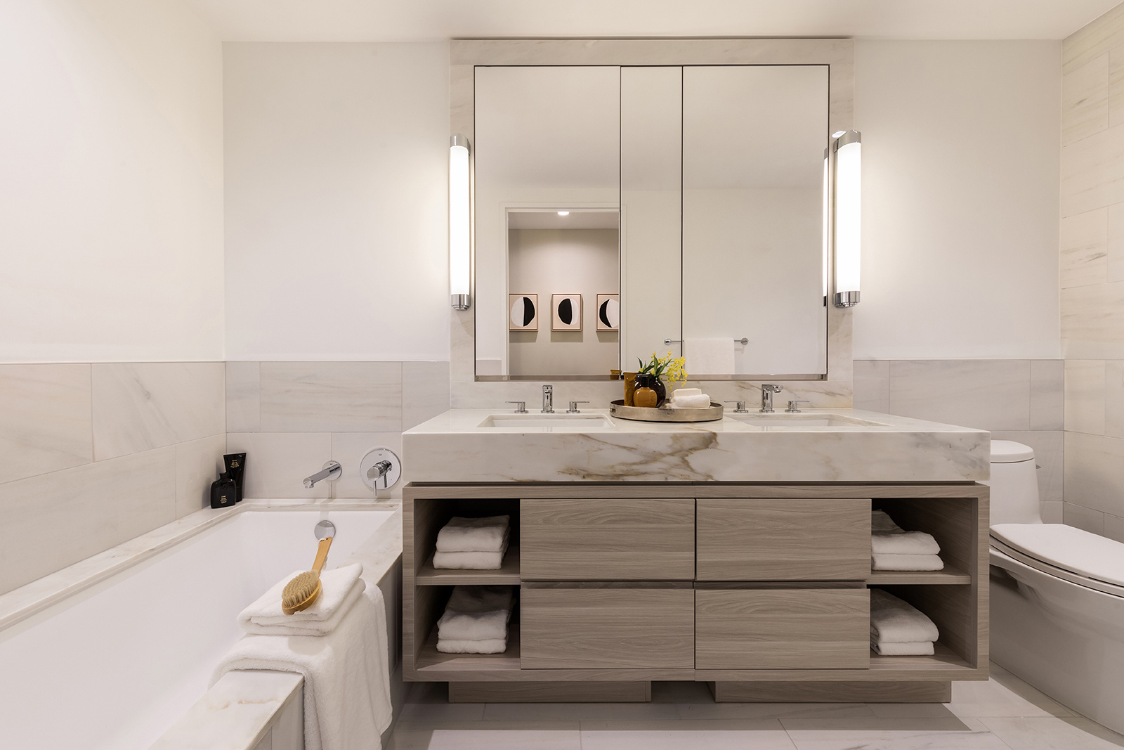 A modern bathroom featuring a large mirror, dual sinks, wooden vanity, white towels, and a bathtub.