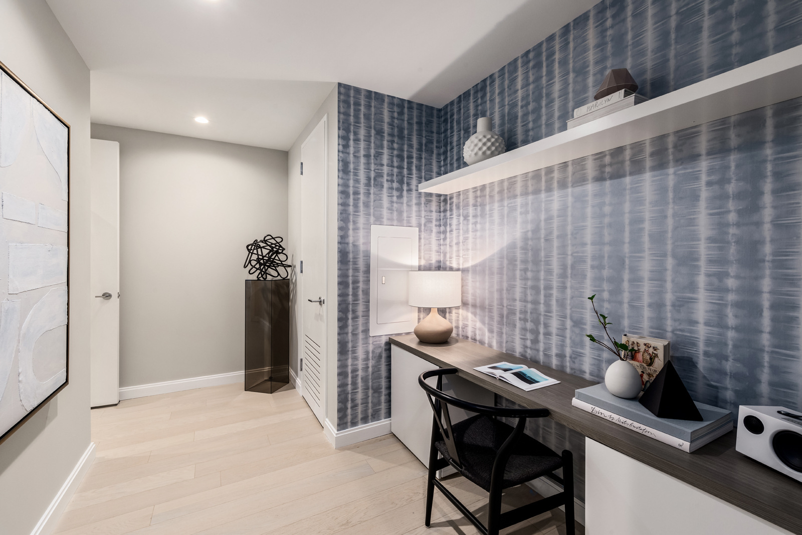 A modern home office nook with a sleek black chair, a tidy desk hosting a few decorative items, and a floating shelf above against a textured blue wallpaper background, creating a serene workspace within a well-lit room.