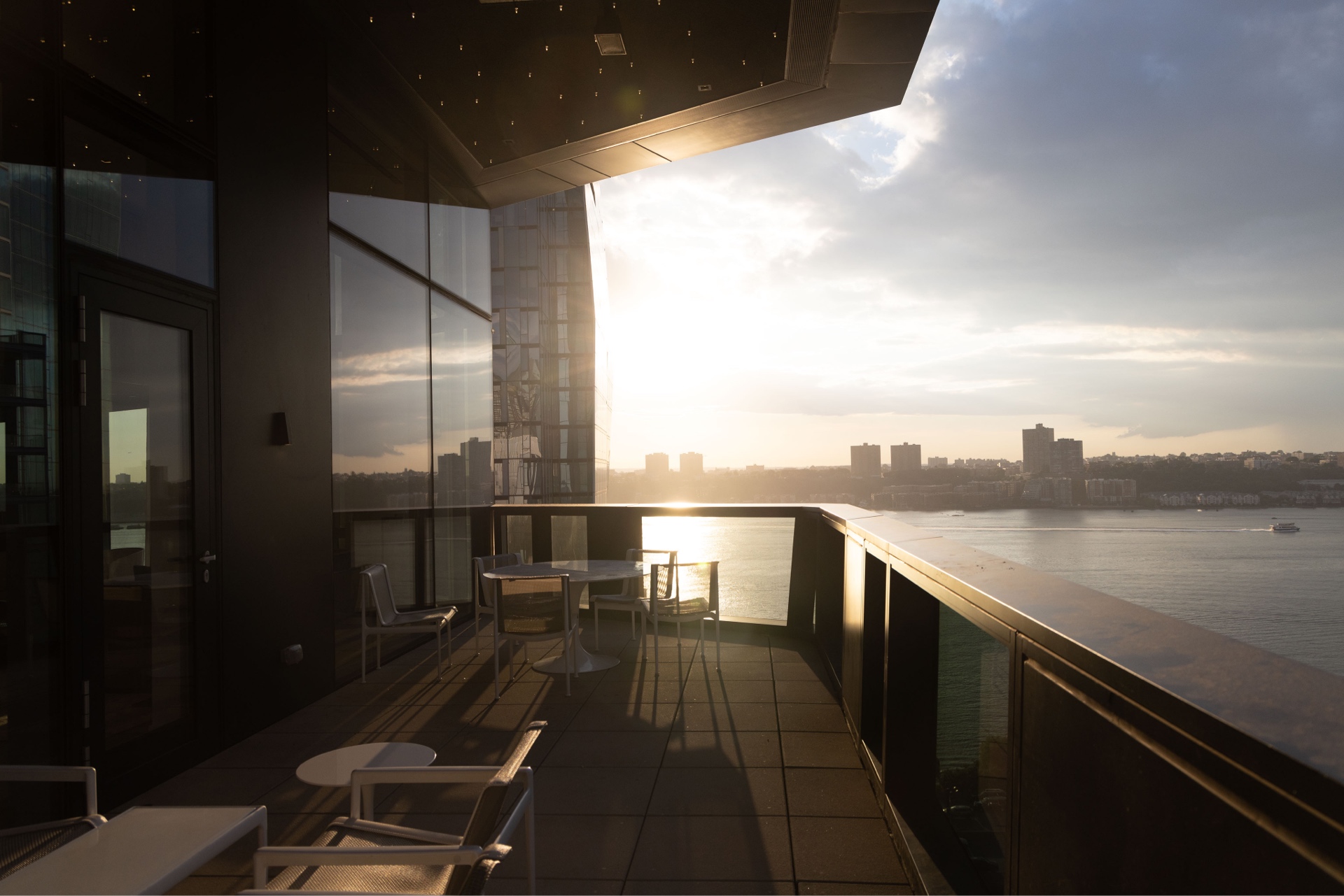 A serene sunset view from a modern balcony overlooking a calm river and a cityscape, with the warm glow of the sun casting reflections on the glass balustrade and creating a tranquil atmosphere.