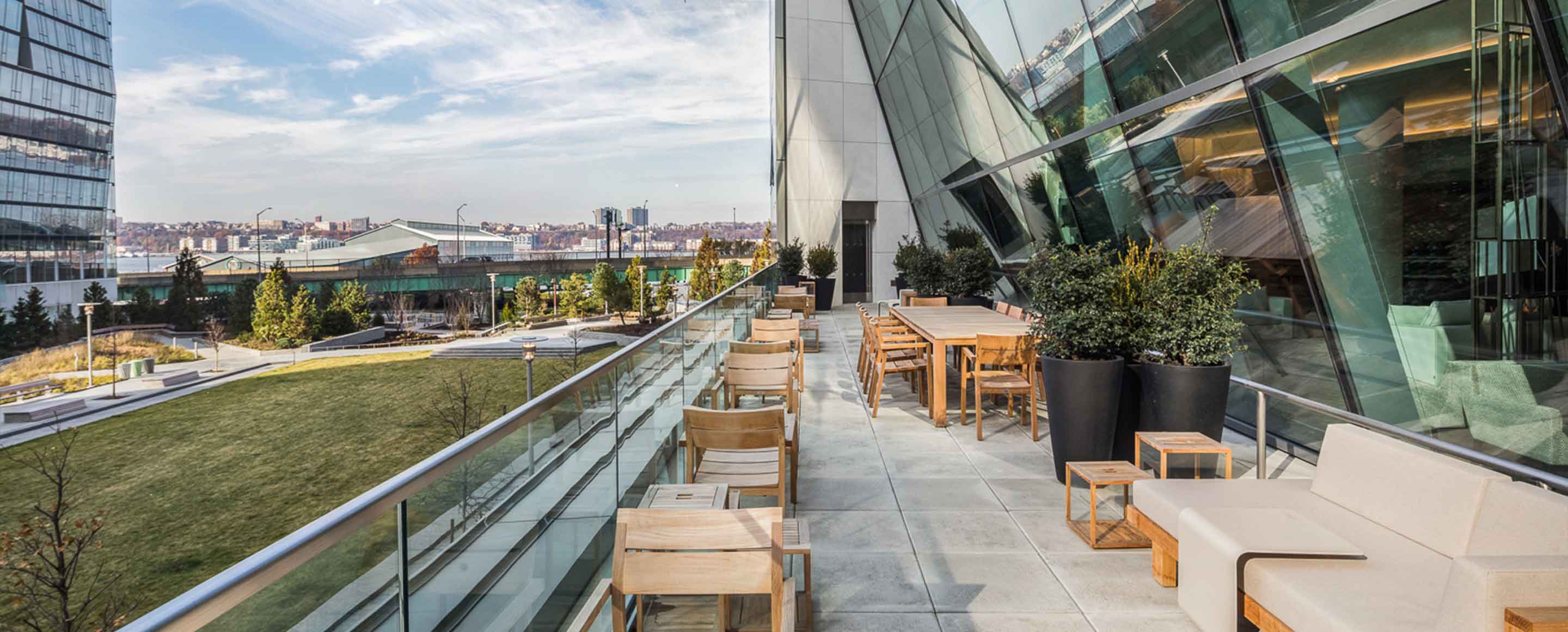 An urban oasis: a sleek terrace with modern wooden furniture overlooking a cityscape, bordered by contemporary glass architecture that reflects the sky and surroundings.