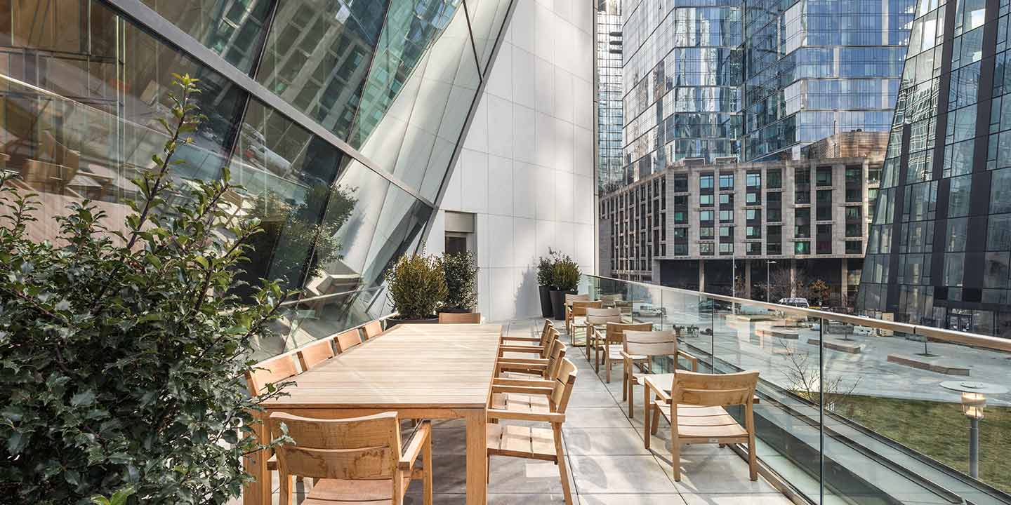 A modern outdoor terrace nestled between high-rise buildings, featuring wooden tables and chairs, green plants, and glass balustrades, offering an urban oasis in a bustling cityscape.
