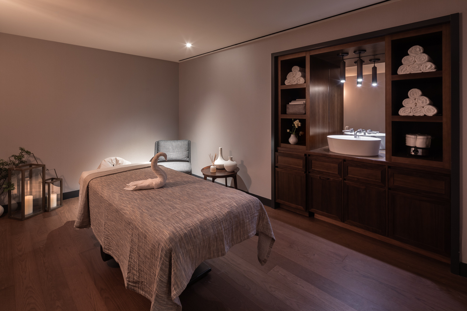 A serene and elegantly decorated massage room with a neatly made treatment table, soft lighting, and a wooden cabinet showcasing neatly folded towels and decorative items.