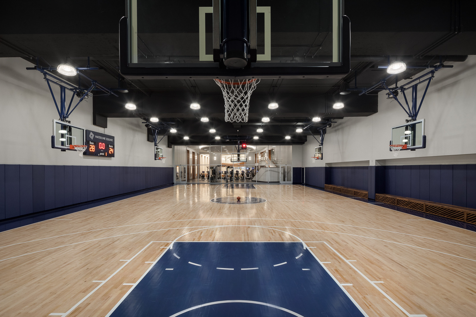An indoor basketball court with a polished wooden floor and multiple hoops, featuring blue padded walls and benches along the sides, with a fitness area visible through the glass doors at the back.