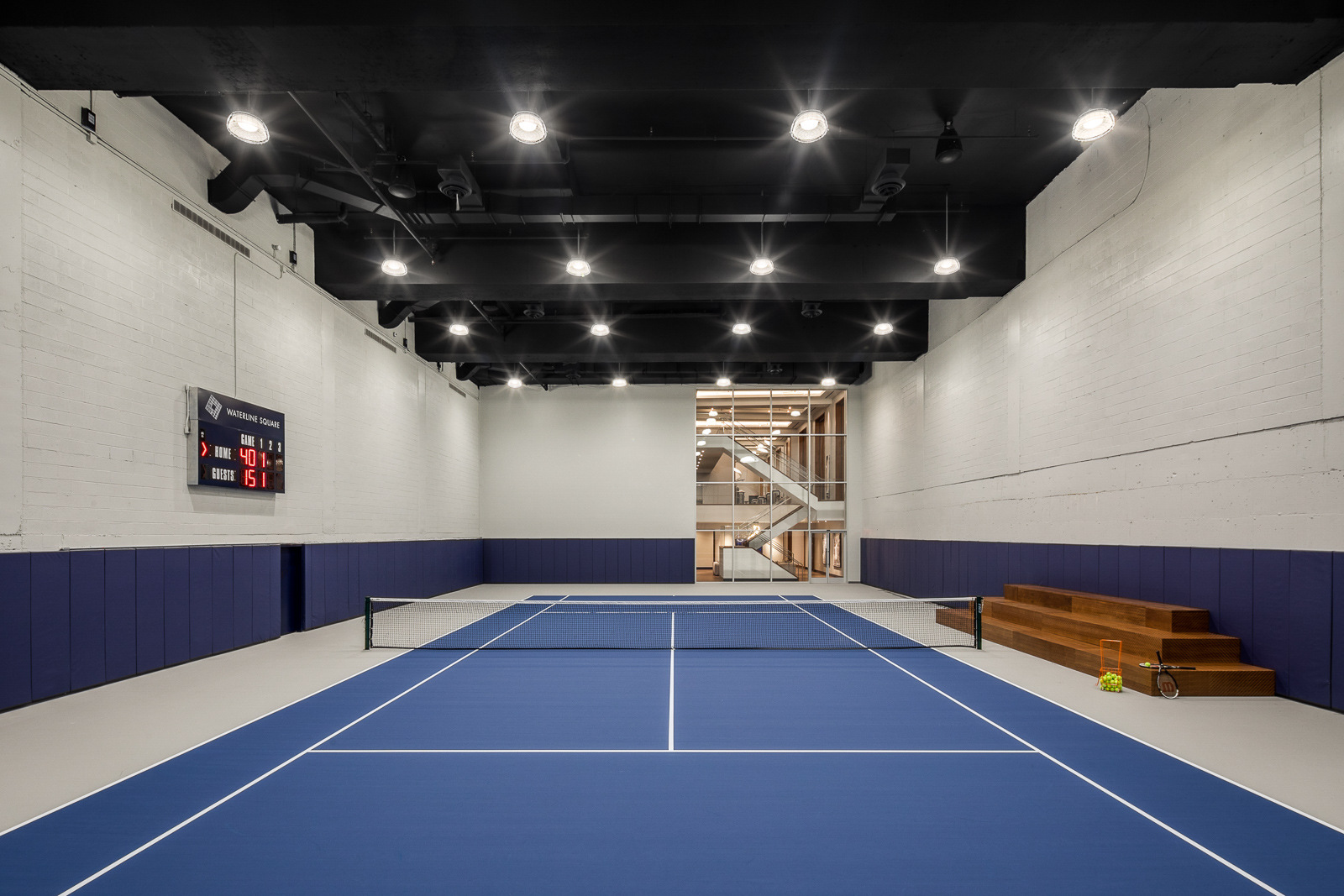 A spacious indoor tennis court with blue flooring, white walls, and elevated spectator seating area, featuring a stairway in the background leading to additional levels.