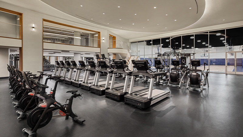 Modern and spacious gym interior with a row of treadmills in the foreground, stationary bikes, and various exercise machines, featuring a mirrored wall and an upper mezzanine level, ready for a workout session.