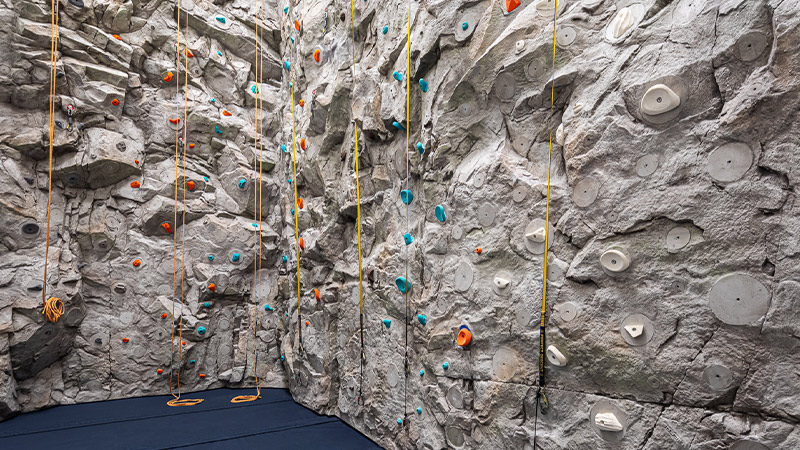 An indoor rock climbing wall equipped with various handholds and footholds, and ropes for safety, set against a textured, grey, artificial rock surface.