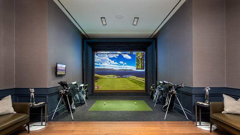 An indoor golf simulator room with a large screen displaying a virtual golf course, flanked by golf equipment and comfortable seating.
