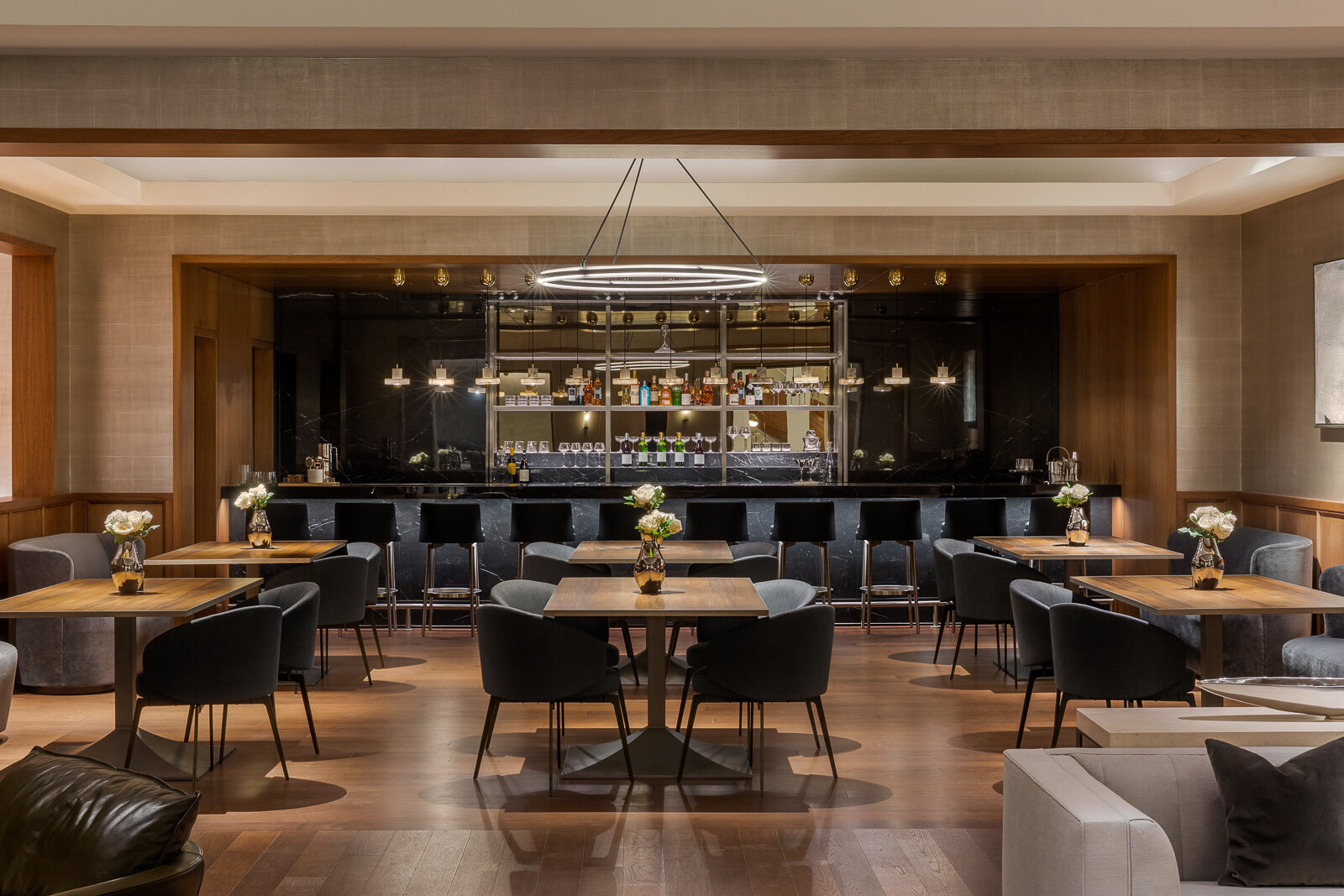 Elegant modern restaurant interior with dark wood tables, comfortable chairs, and a well-stocked bar illuminated by soft ambient lighting.