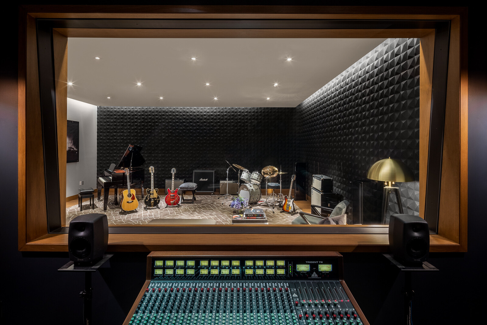 A modern and well-equipped music recording studio, viewed from the control room through a large window, featuring a selection of guitars, a drum set, keyboards, and soundproofing on the walls.