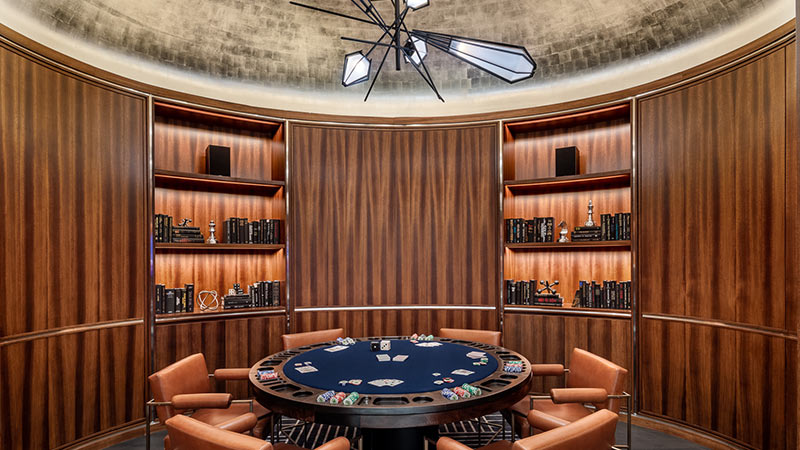 A luxurious, circular room with wood-paneled walls, elegantly lined with bookshelves and decorative items, centered around a sophisticated poker table under a modern light fixture.