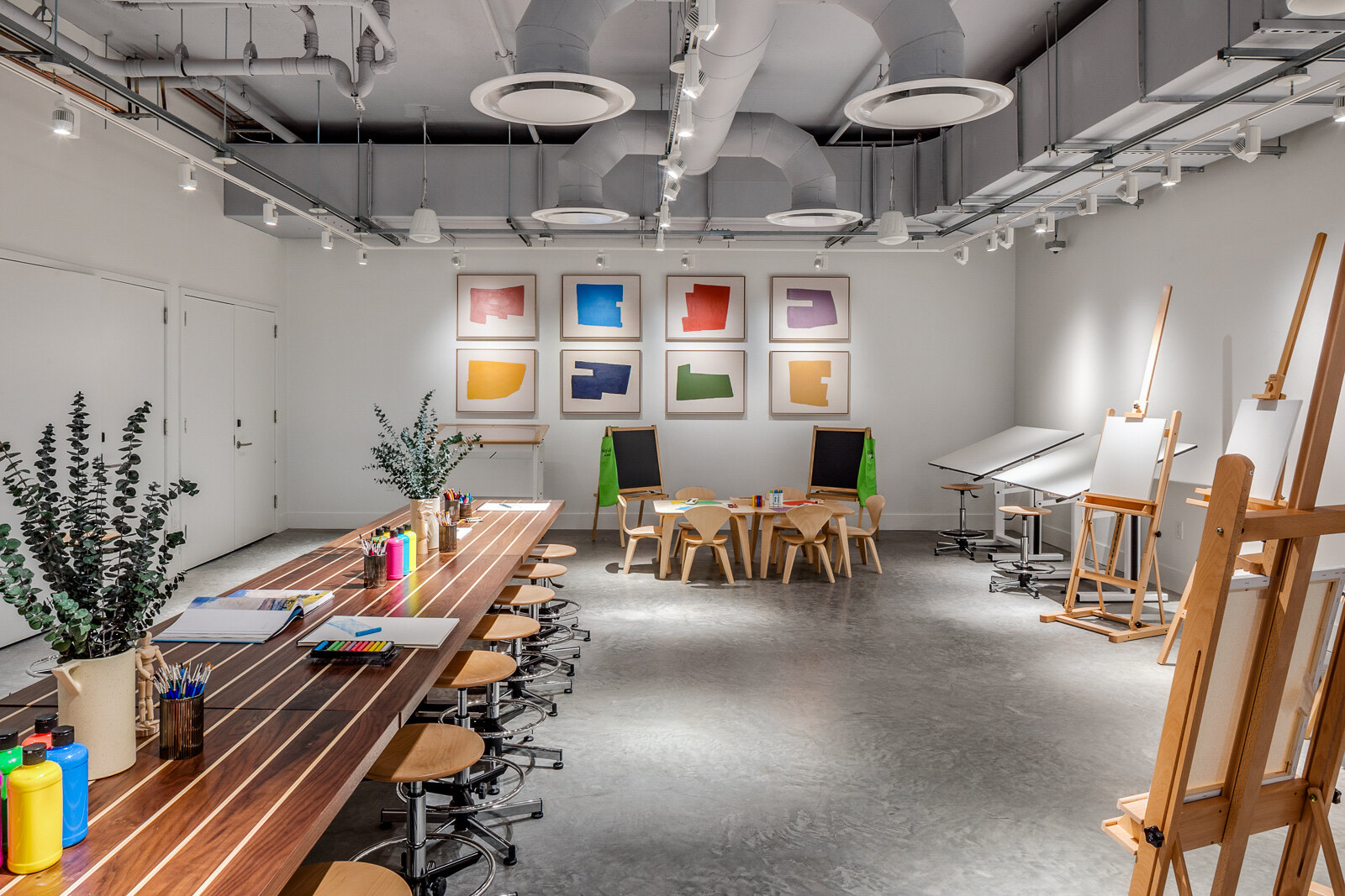 A modern and bright art studio with easels, a long worktable with art supplies, and colorful artwork displayed on the wall.