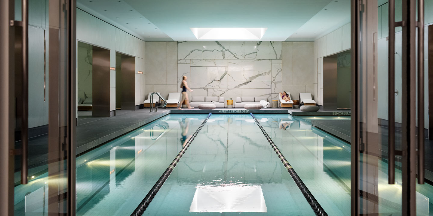 An indoor swimming pool in a luxurious, modern space with clean lines, reflected in the still water, with a person walking alongside the pool.