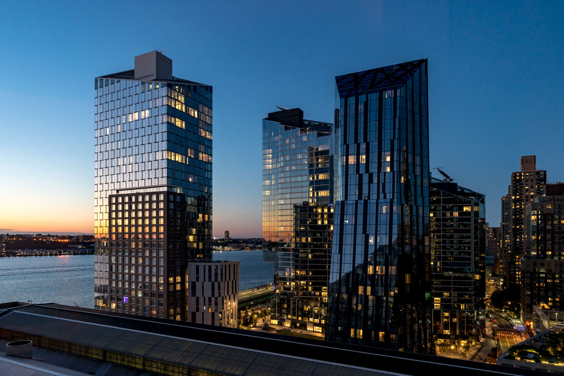 Modern skyscrapers dominate the cityscape against the backdrop of a twilight sky, with indoor lights beginning to twinkle as evening falls over the urban waterfront.