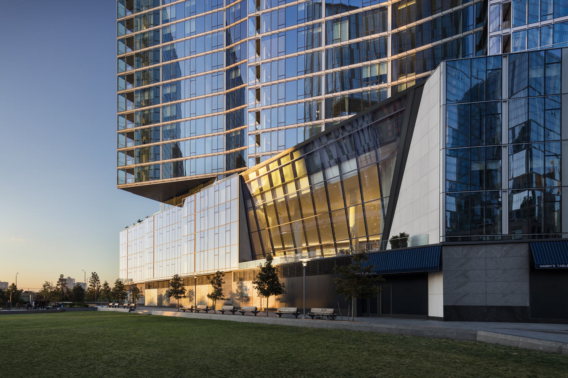 Modern architecture meets golden hour: a sleek glass building basks in the warm glow of the setting sun, reflecting the clear blue sky, as shadows stretch across the manicured lawn.