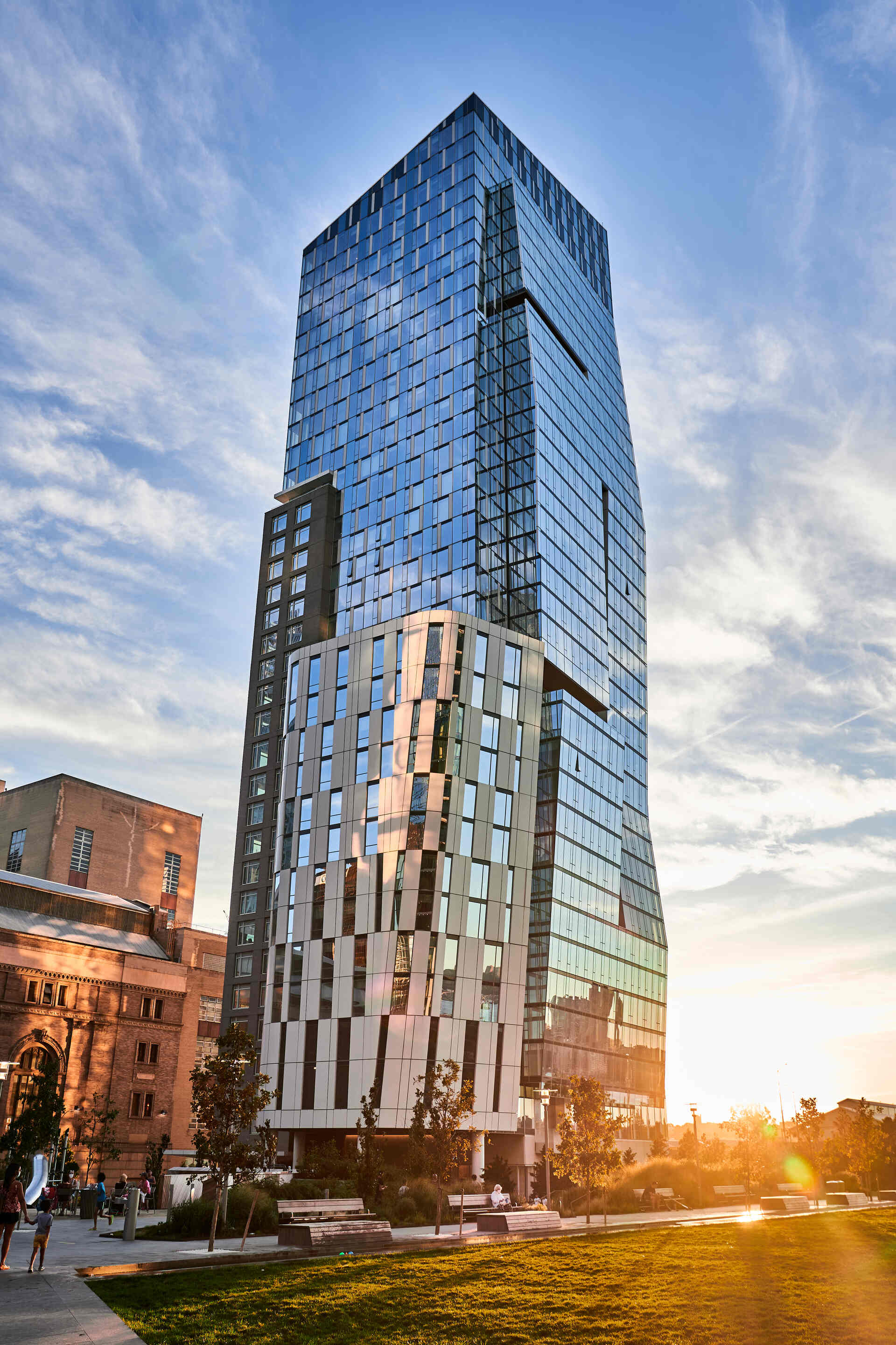 Modern glass building towering into the evening sky flanked by warm sunlight and an older structure to the left, with people enjoying a stroll in the urban park below.