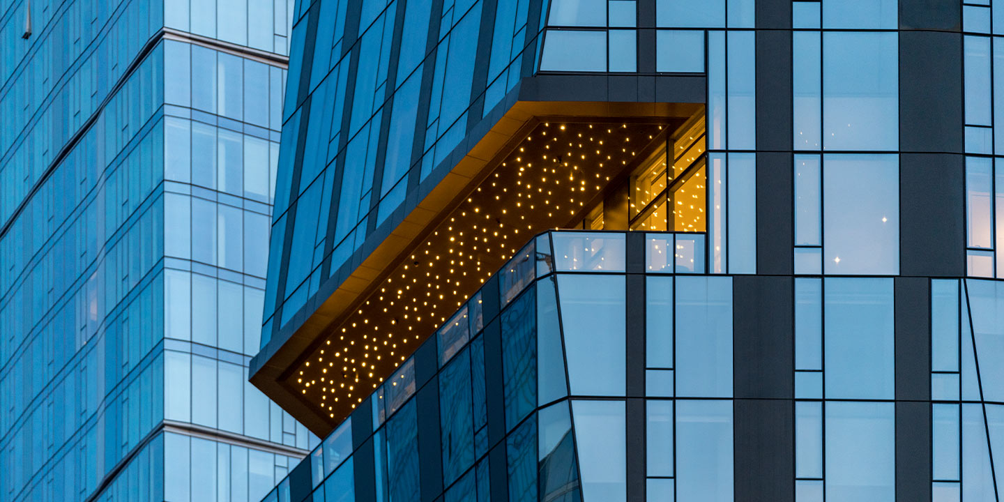 Twilight hues envelop a modern glass building, where a uniquely lit balcony juts out, offering a warm glow amidst the cool blue architecture.