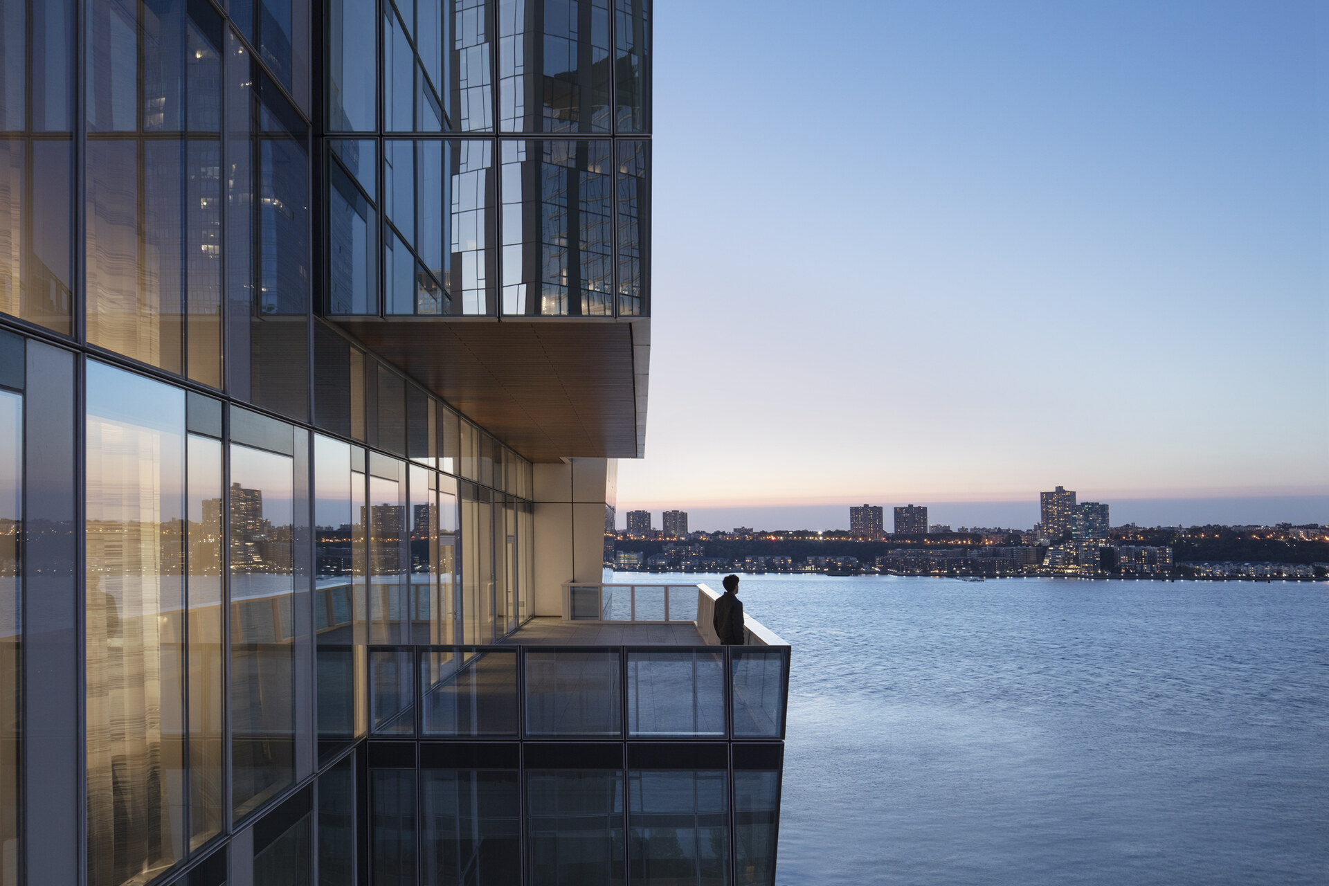 Contemplation at dusk: a lone figure stands on a modern balcony overlooking a tranquil waterfront cityscape as the evening light fades.