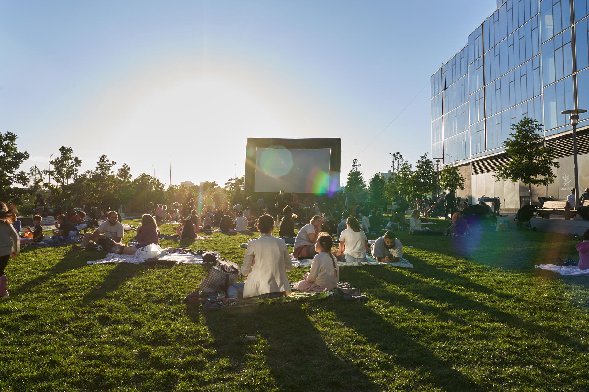 Outdoor community movie night on a sunny evening with people sitting on the grass enjoying a film on a large inflatable screen.