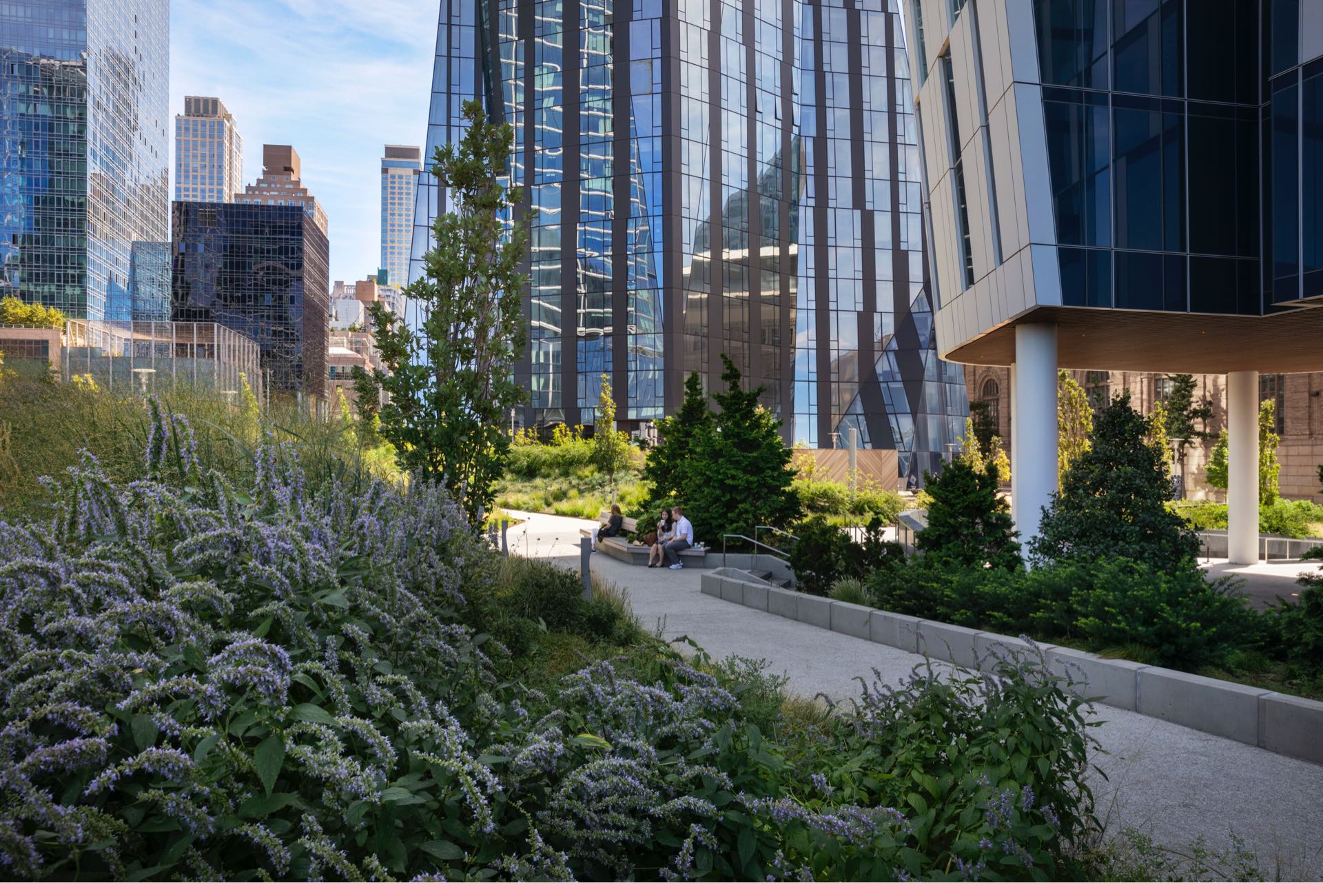 A tranquil urban garden nestled among modern high-rise buildings, with people enjoying a sunny day amidst lush greenery and blooming purple flowers.