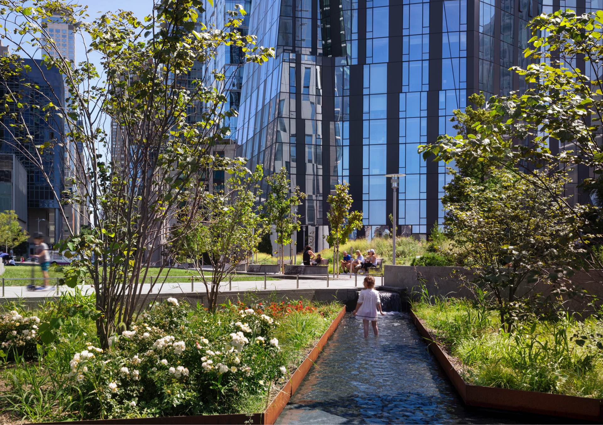 A young child walks alongside a narrow water feature in an urban park, with lush greenery and towering reflective skyscrapers in the background.