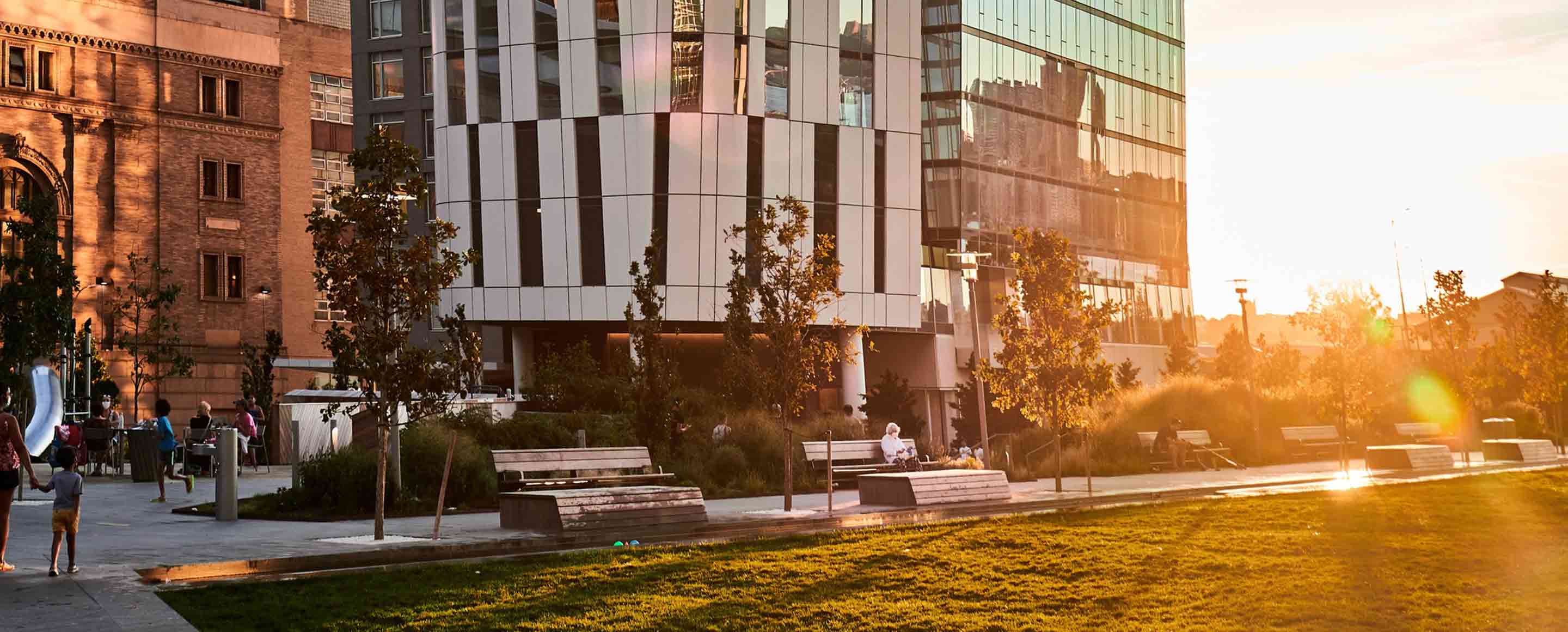 A peaceful urban park at sunset with people relaxing on benches and walking, enjoying the warm glow of the setting sun against modern buildings.