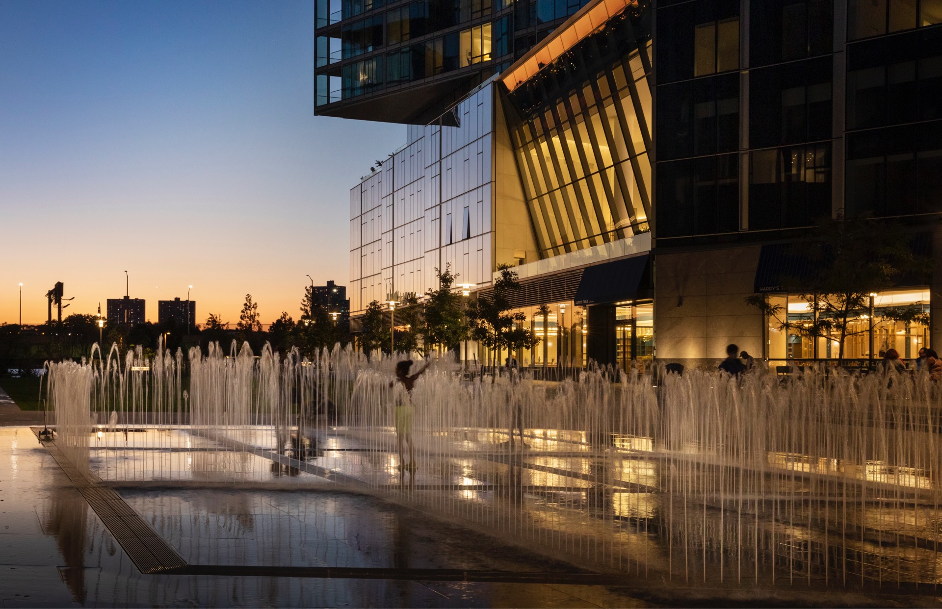 Urban twilight at a modern plaza with illuminated water fountains and sleek architectural buildings, where silhouettes of people enjoy a serene evening outdoors.