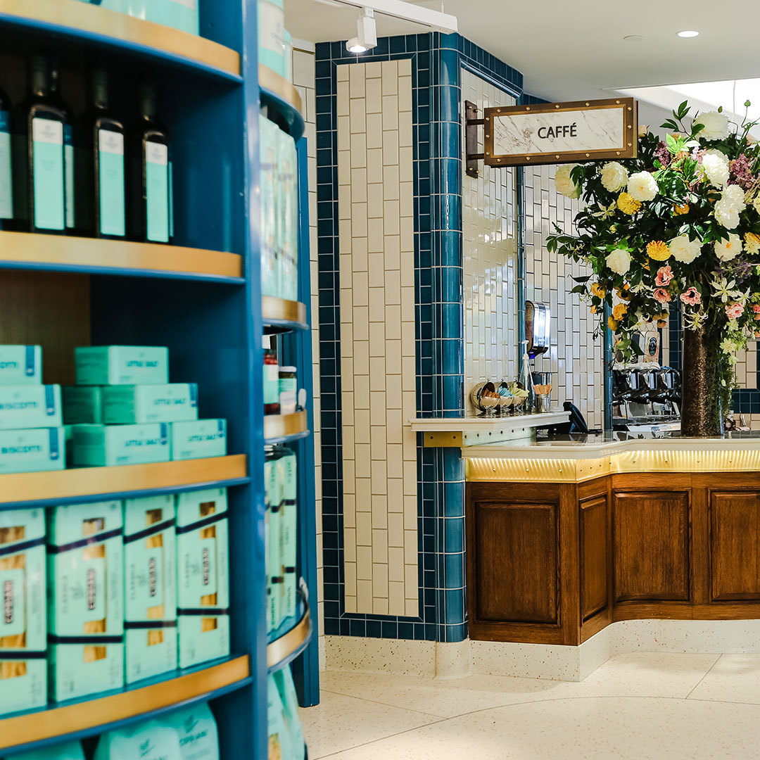 An inviting café corner with a vintage aesthetic, showcasing elegant tiling, wooden counters, and an array of neatly arranged products, complemented by a vibrant floral display.