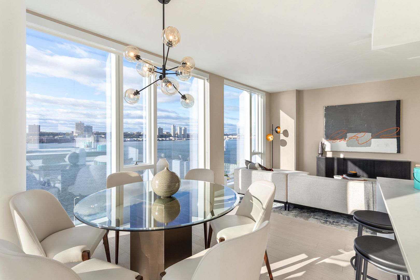 Bright and modern dining area with a glass table and leather chairs, boasting an elegant chandelier and panoramic views of the waterfront through floor-to-ceiling windows.